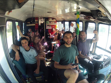 Eine Gruppe von Freunden sitzt fröhlich in einem dekorierten Bus, der mit Büchern und persönlichen Gegenständen gefüllt ist, lächelt, winkt und ist bereit für ein lustiges Roadtrip-Abenteuer.