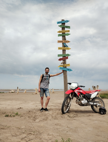Ein Mann steht neben einem Wegweiser mit Städtenamen und Entfernungen, neben einem roten Motorrad in einer Sandlandschaft unter einem bewölkten Himmel und wirkt abenteuerlustig.