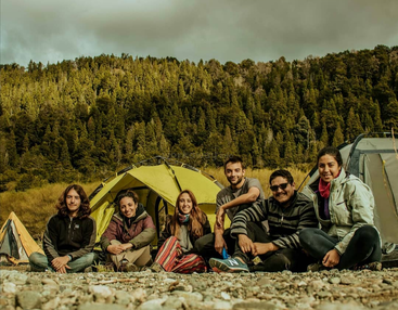 Sechs Freunde sitzen lächelnd auf dem felsigen Boden vor ihren Zelten, umgeben von üppig grünem Wald unter einem wolkenverhangenen Himmel, und genießen ein gemeinsames Camping-Abenteuer im Freien.