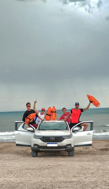Vier junge Männer, möglicherweise Rettungsschwimmer, posieren fröhlich mit orangefarbenen Rettungsbojen und einem weißen Fiat-Auto an einem Sandstrand mit dem Meer im Hintergrund.