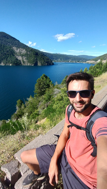 Ein Mann mit Sonnenbrille und Rucksack lächelt für ein Selfie mit einem wunderschönen blauen See, grünen Bergen und einem klaren, sonnigen Himmel im Hintergrund.