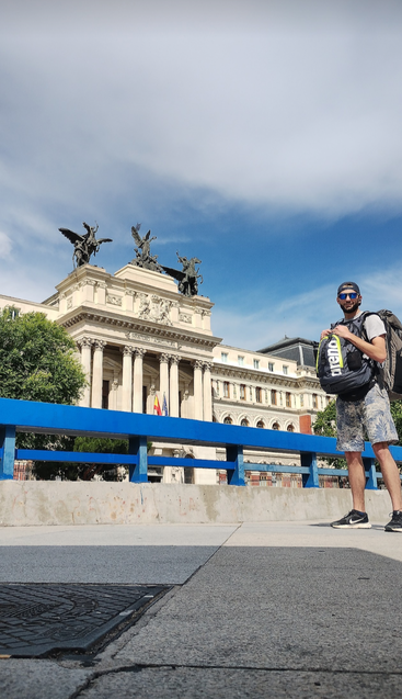 Ein Mann mit einem Rucksack steht auf dem Bürgersteig vor einem verzierten historischen Gebäude mit Statuen auf dem Dach, unter einem hellen, teilweise bewölkten Himmel.