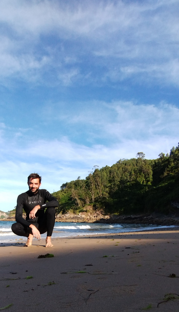 Ein Mann im Neoprenanzug hockt an einem Sandstrand mit Seegras, neben sanften Wellen, üppig grünen Hügeln und einem strahlend blauen Himmel mit Wolken.