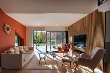 A bright, modern living room features a beige sofa, cozy armchairs, wooden accents, large glass doors opening to a garden, and a TV mounted on the wall.