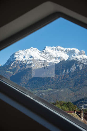 A picturesque view of snow-capped mountains framed by a window, green hills below, rooftops in the foreground, under a clear blue sky, scenic and serene.