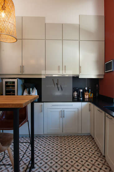 Modern kitchen with white cabinets, black countertops, patterned tile floor, wooden island, leather and wooden stools, coffee machine, food processor, and stylish rattan pendant light.