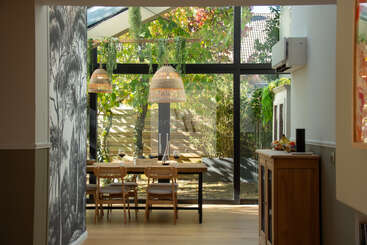 A bright dining area with large glass windows overlooks a lush garden. Wooden furniture, hanging lamps, and plants create a cozy, natural, and inviting atmosphere.