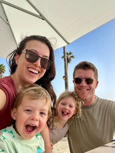 A smiling family of four, two adults and two young children, take a cheerful selfie at the beach under a large umbrella, with palm trees behind them.