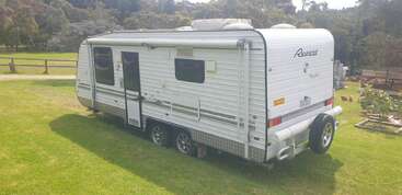 A white Regent caravan with dual axles is parked on a grassy area, surrounded by trees and wooden fences, under a bright, sunny sky.