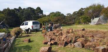 A white van is parked on grass, with a person working nearby. Cut logs and piles of uprooted plants are scattered across a spacious, green area.