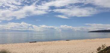 A serene sandy beach stretches along calm, reflective water under a vast sky filled with fluffy clouds. Distant hills and greenery complete this peaceful coastal scene.