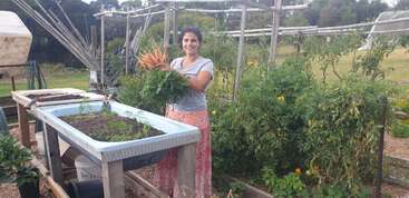 A woman stands smiling in a thriving garden, holding a large bundle of freshly harvested carrots, surrounded by lush green plants and raised garden beds.