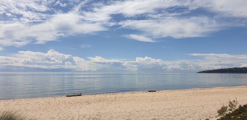 Ein ruhiger Sandstrand erstreckt sich entlang des ruhigen, spiegelnden Wassers unter einem weiten Himmel voller flauschiger Wolken. Entfernte Hügel und Grünflächen vervollständigen diese friedliche Küstenszene.