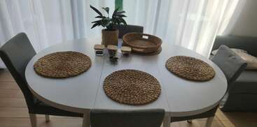 Uma mesa de jantar oval branca com quatro cadeiras cinza é colocada com jogos americanos de tecido. Uma planta, cestas e condimentos decoram a mesa sob cortinas transparentes.