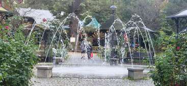 Uma cena encantadora de parque com uma fonte circular com arcos de água, plantas verdes exuberantes, bancos, caminhos de paralelepípedos e duas pessoas caminhando ao fundo.