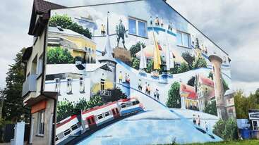 Um mural vibrante em um edifício mostra uma cena animada da cidade com trens, veleiros, pessoas, vegetação, pontos de referência notáveis e edifícios coloridos, todos se misturando perfeitamente.