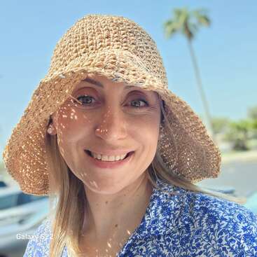 Uma mulher usando um chapéu de sol de palha e uma camisa com estampa azul sorri ao ar livre sob o sol forte, com palmeiras e céu azul ao fundo.