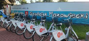 L'image montre une rangée de vélos de location garés dans une station de vélos en libre-service. Derrière eux, une peinture murale colorée indique en caractères gras "Kocham Grodzisk Mazowiecki".