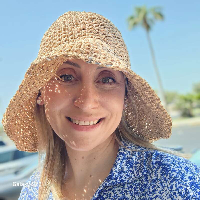 Uma mulher usando um chapéu de sol de palha e uma camisa com estampa azul sorri ao ar livre sob o sol forte, com palmeiras e céu azul ao fundo.