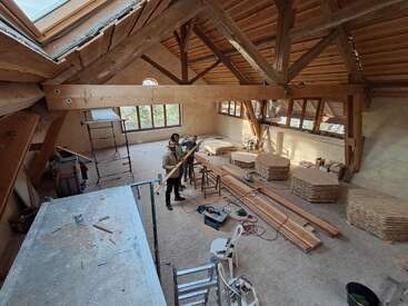Two people are working in a spacious, wooden-beamed room under construction, surrounded by tools, ladders, wood planks, and large piles of cut wooden boards.