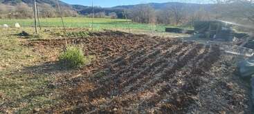 A freshly tilled garden plot with rows of soil, surrounded by a scenic rural landscape, green fields, trees, and distant hills under a clear blue sky.