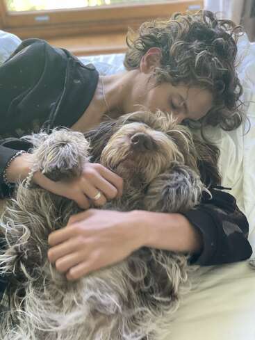 Una persona con el pelo rizado acaricia con cariño a un perro peludo en una cama; ambos parecen relajados y felices, disfrutando juntos de la tranquilidad de una habitación iluminada por el sol.