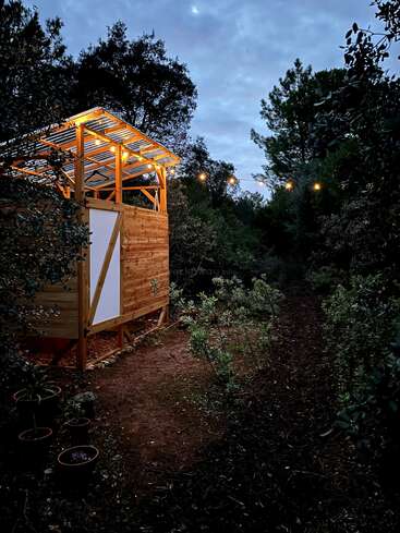 La imagen muestra una estructura de madera con tejado metálico, situada en una zona boscosa, con luces de cuerda y una puerta blanca, rodeada de árboles y arbustos.