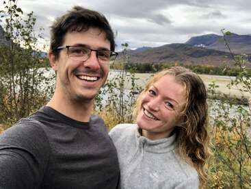 L'image représente un jeune couple prenant un selfie devant un lac serein, entouré de montagnes et d'arbres sous un ciel couvert.