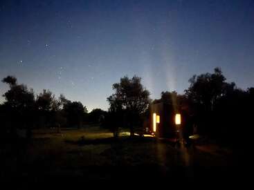 La imagen representa una serena escena nocturna en la que aparece un pequeño edificio con ventanas iluminadas, situado en medio de una zona de césped rodeada de árboles bajo un cielo estrellado.