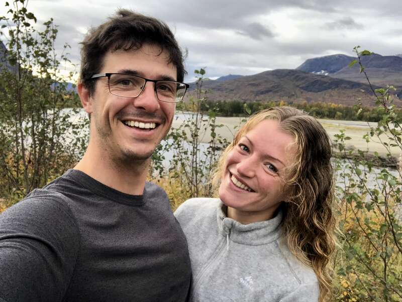 La imagen muestra a una joven pareja tomándose un selfie frente a un lago sereno, rodeado de montañas y árboles bajo un cielo nublado.