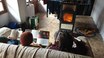 Two people are reading on a cozy couch near a glowing wood stove. Children's toys and laundry hang nearby, creating a warm, homely, and relaxed atmosphere.
