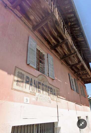 This image shows an old pink building with weathered shutters, a wooden balcony, and a faded sign reading "PANETTERIA." Sunlight softly illuminates the charming, rustic exterior.