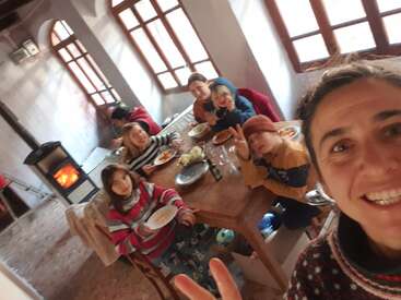 A smiling woman takes a selfie with children around a wooden table, enjoying a meal together in a cozy room with a wood stove and large windows.