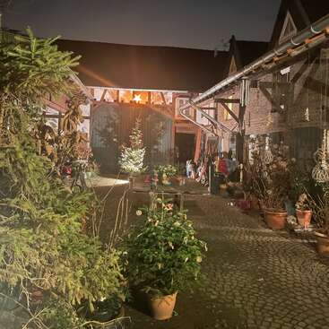 The image depicts a serene courtyard at night, featuring a brick building with a sloping roof, surrounded by potted plants and trees, and illuminated by a warm light source.