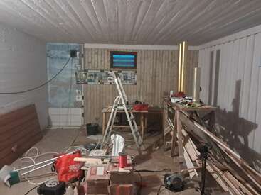 This image shows a cluttered workshop or renovation area with tools, wooden planks, a step ladder, scattered materials, and unfinished walls, indicating ongoing construction or remodeling work.