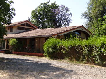 L'image représente une maison à deux étages avec un toit brun et un bardage beige, entourée d'arbres et de buissons, avec une allée en gravier au premier plan.
