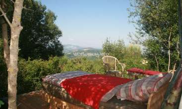 The image depicts a serene outdoor setting with a bed, chair, and table situated on a wooden deck, surrounded by lush trees and a breathtaking view of the countryside.
