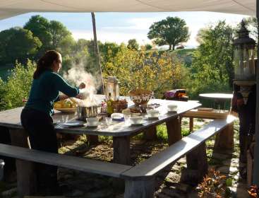 The image depicts a woman cooking on a large outdoor table, surrounded by trees and a serene landscape. The scene exudes a sense of tranquility and natural beauty.