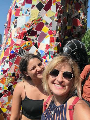 Zwei Frauen lächeln strahlend vor einer bunten Mosaikwand im Sonnenlicht. Eine von ihnen trägt eine Sonnenbrille. Beide wirken glücklich und genießen die lebendige, künstlerische Umgebung im Freien.