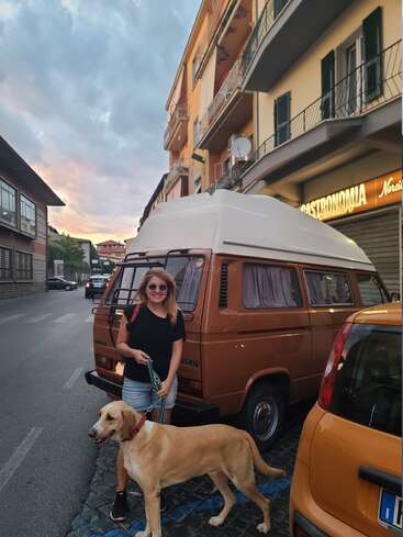Eine Frau mit Sonnenbrille und schwarzem Hemd steht lächelnd mit einem großen hellbraunen Hund an der Leine neben einem braunen Lieferwagen auf einer Stadtstraße bei Sonnenuntergang.