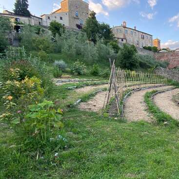 Um jardim em terraços com treliças de bambu e plantas verdes fica numa encosta, com charmosos edifícios de pedra ao fundo, sob um céu azul parcialmente nublado. Uma cena tranquila.
