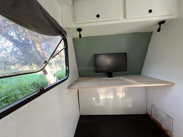 Ein minimalistischer Arbeitsplatz in einem kleinen Raum oder einem Kleinbus verfügt über ein großes Fenster, einen Schreibtisch aus Holz, einen Computermonitor, eine grüne Akzentwand, Stauraum an der Decke und einen Blick auf die grüne Umgebung draußen.