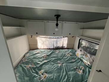 Ein gemütliches Schlafzimmer im Wohnmobil verfügt über ein Doppelbett mit einer Bettdecke mit Gänseblümchenmuster, Hängeschränke, weiße Wände, ein Fenster mit Vorhang, Ablageflächen und eine einzelne Glühbirne.