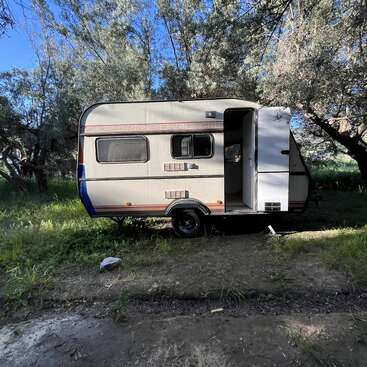 Une petite caravane est garée dans un coin ombragé et verdoyant, au milieu de grands arbres. La porte est ouverte ; la lumière du soleil filtre à travers les feuilles, créant un cadre paisible et naturel.