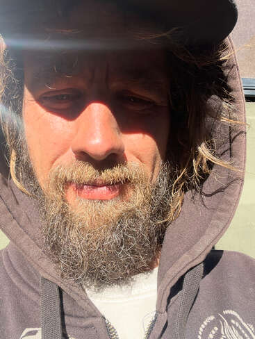 A man with a full, scruffy beard and wavy hair wears a hoodie and hat. Sunlight creates shadows across his face, as he squints slightly.