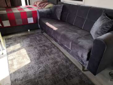 Ein gemütlicher Wohnbereich mit einem großen grauen Samtsofa, zwei passenden Kissen, einem weichen Teppich und einem Bett, das mit einer rot karierten Decke bedeckt ist.