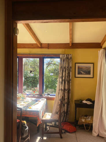 Ein sonniges Zimmer mit Holzbalken, gelben Wänden und einem großen Fenster. Eine farbenfrohe Tischdecke bedeckt einen Tisch mit Stühlen, Pflanzen, Büchern und einfallendem Sonnenlicht. Gemütliche Atmosphäre.