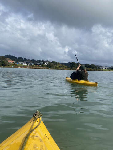 Eine Person paddelt in einem gelben Kajak auf ruhigem Wasser unter einem bewölkten Himmel. Das Foto wurde von einem anderen Kajak aus aufgenommen, und an der Küste sind in der Ferne Häuser zu sehen.