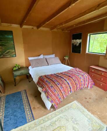 Dieses gemütliche Schlafzimmer hat ein einfaches Bett mit braunen Kissen und einer bunten Decke, Holzmöbel, bunte Teppiche, Zimmerpflanzen und gerahmte Kunstwerke an erdfarbenen Wänden.