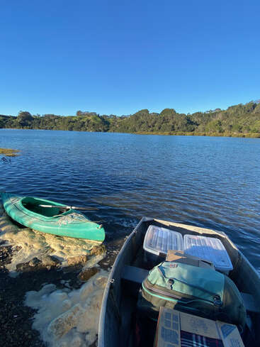 Ein ruhiger See unter strahlend blauem Himmel, ein grünes Kajak schwimmt in der Nähe des Ufers und ein Boot ist mit Ausrüstung beladen, umgeben von üppig grünen Hügeln.
